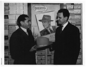 Photograph of Max Goldfine and Homer Stephenson holding a hat inside of Goldfine's department store. An advertisement for Resistol hats with a picture of Errol Flynn is on the wall behind them.